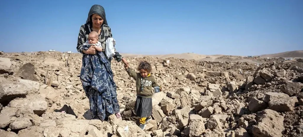 امرأة أفغانية تحمل طفلها وتسير مع طفلة وسط منطقة مدمرة